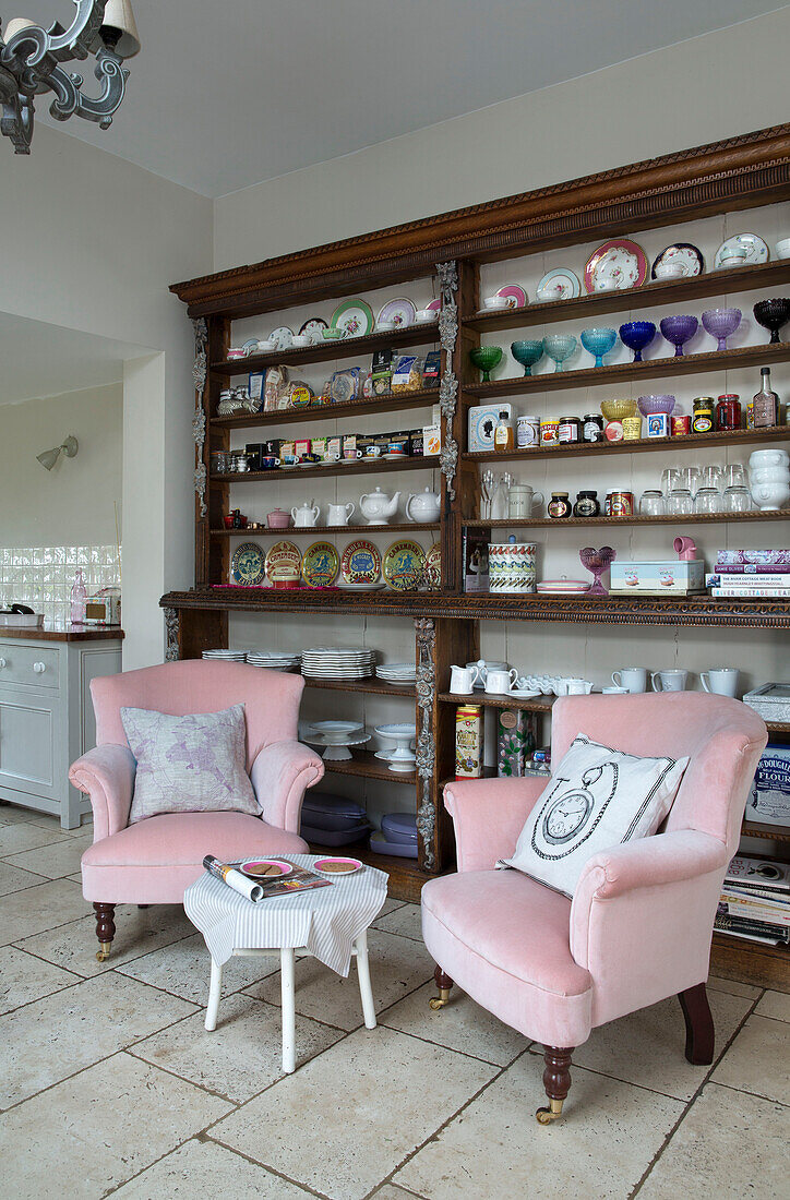 Gläser und Geschirr auf Holzregalen mit zwei rosa Sesseln in einer Küche in Sussex UK