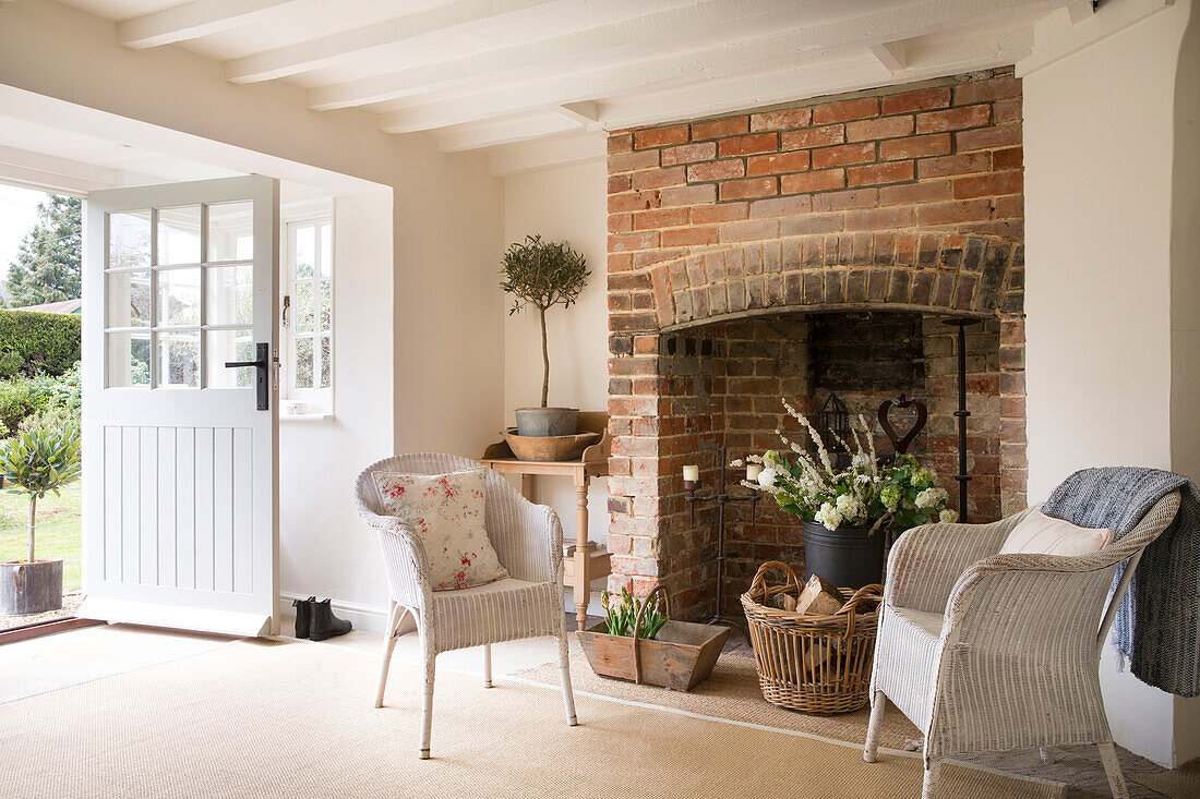 Pair of wicker armchairs at brick fireplace with open back door in East Dean farmhouse  West Sussex  UK