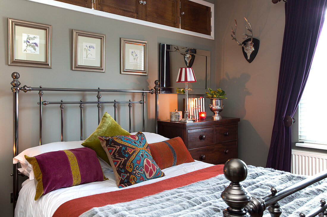 Gemusterte Kissen auf schmiedeeisernem Bett mit gerahmten Kunstwerken und Schrankablage in einem Schlafzimmer in Surrey, England UK