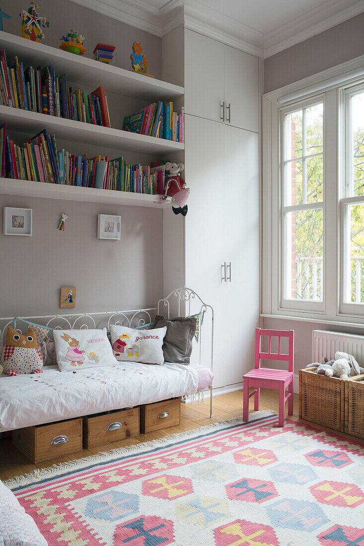 Bücherregal über dem Tagesbett mit kleinem rosa Stuhl im Mädchenzimmer einer Familie im Südwesten Londons UK