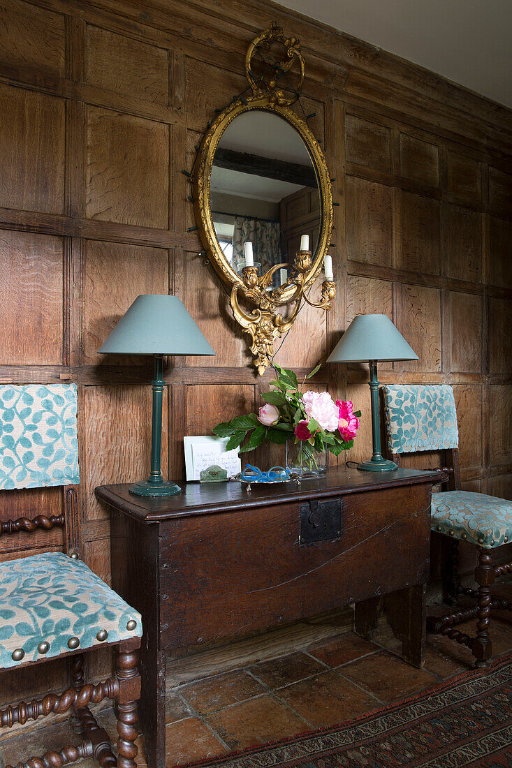 Circular gilt mirror above table with pair of lamps and upholstered chairs in panelled Kent farmhouse UK