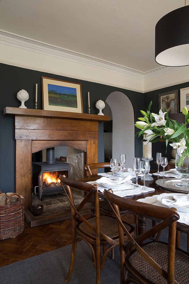 Lit woodburning stove in dining room with wooden table and chairs Gloucestershire home England UK