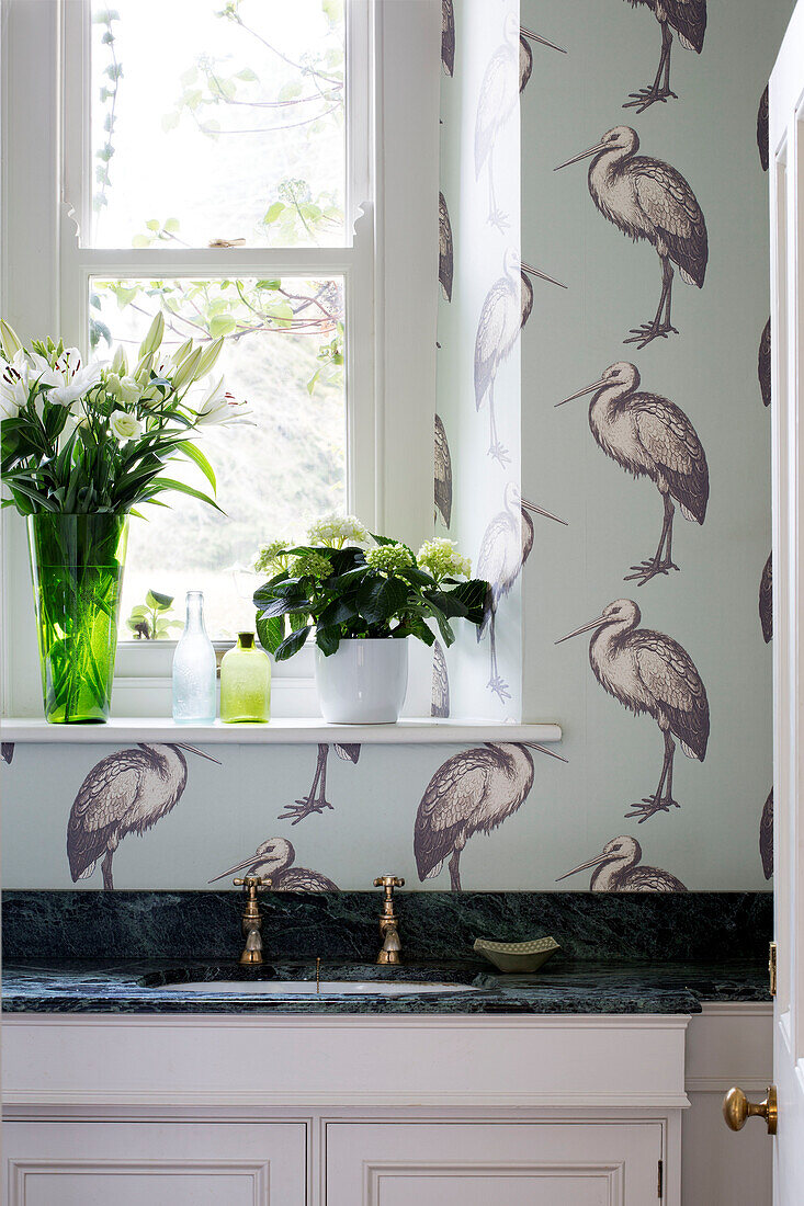 Flowers on windowsill with patterned stork wallpaper above sink in Gloucestershire home England UK