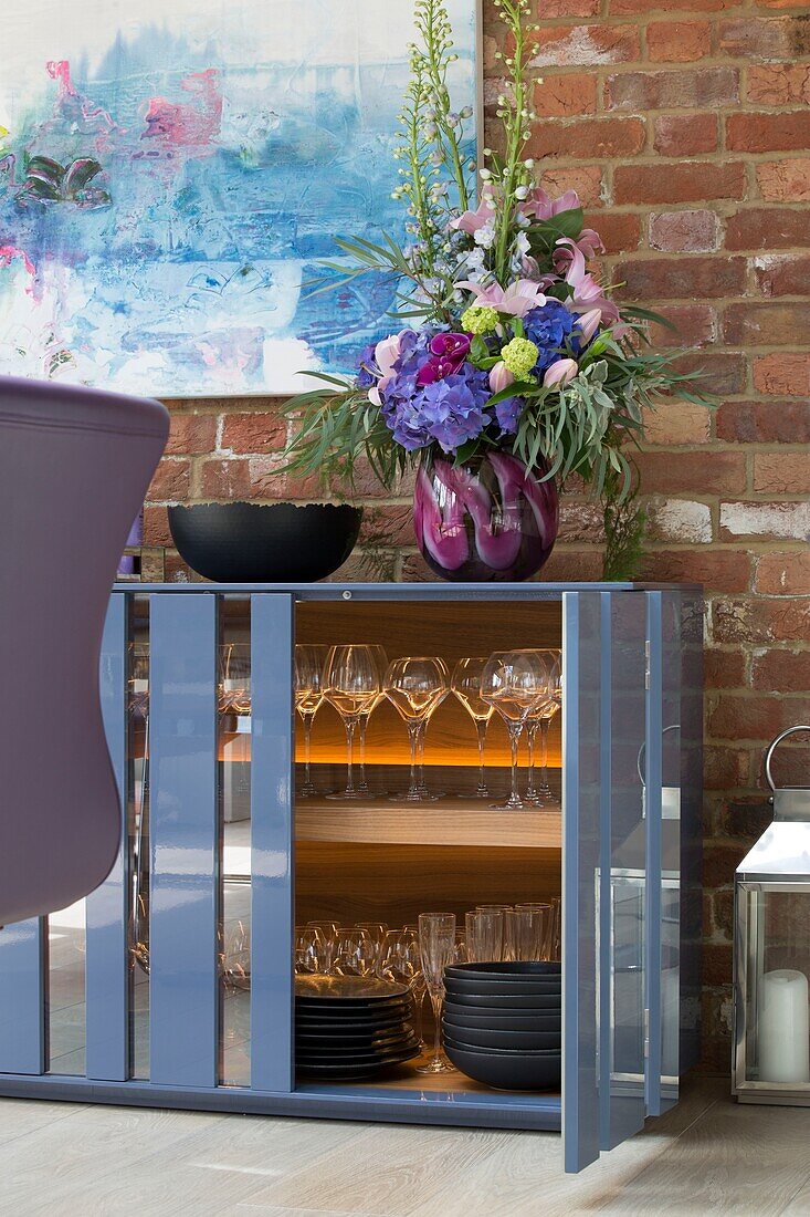 Gläser und Geschirr in blauem Sideboard mit freiliegender Ziegelwand in einem Haus in Sussex, England, UK