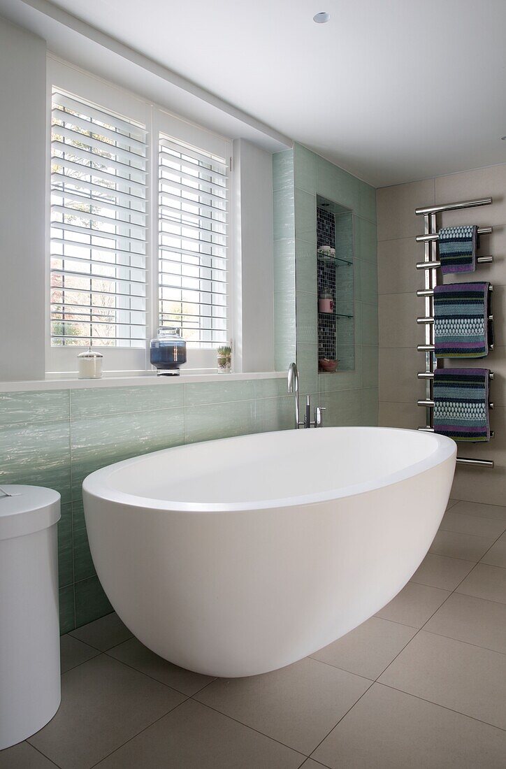 Freistehende Badewanne unter dem Fenster mit Jalousien und wandmontiertem Handtuchhalter in einem Haus in Sussex, England UK