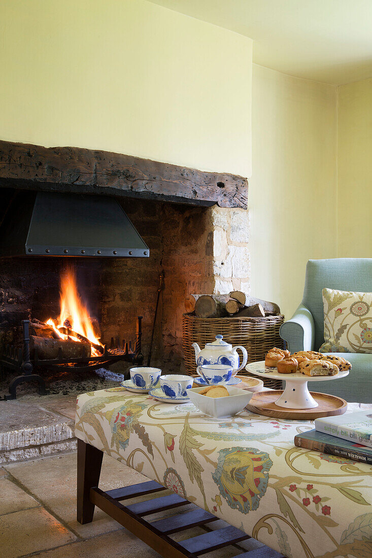 Teaset and cakes on patterned ottoman at lit fireside in Gloucestershire farmhouse England UK