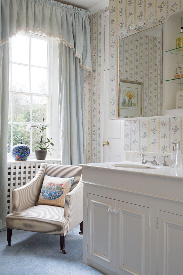 Hellblaue Vorhänge mit Schabracke und Stuhl neben einem Waschbecken mit Spiegelschrank in einem Bauernhaus in Gloucestershire, England, UK