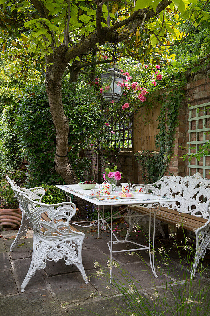 Weißes schmiedeeisernes Sitzmöbel mit Tisch auf der Terrasse unter einem Baum im Garten eines Londoner Hauses, Großbritannien
