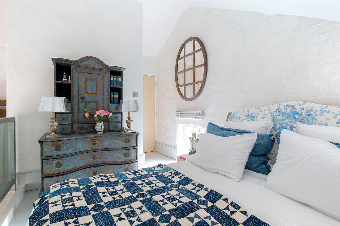 Blue patchwork quilt with ageing dresser and circular mirror in bedroom of South London schoolhouse conversion UK