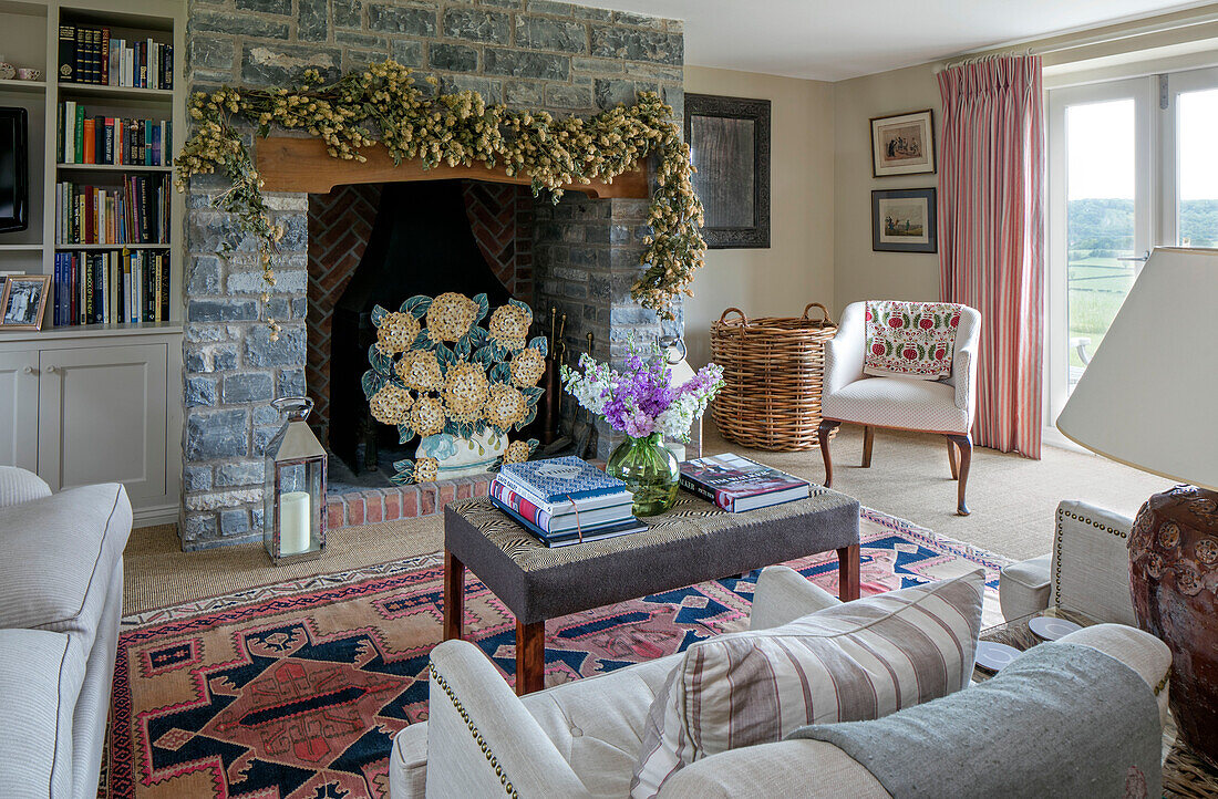 Flower garland above stone fireplace with bookshelf and log basket in living room of Somerset farmhouse UK
