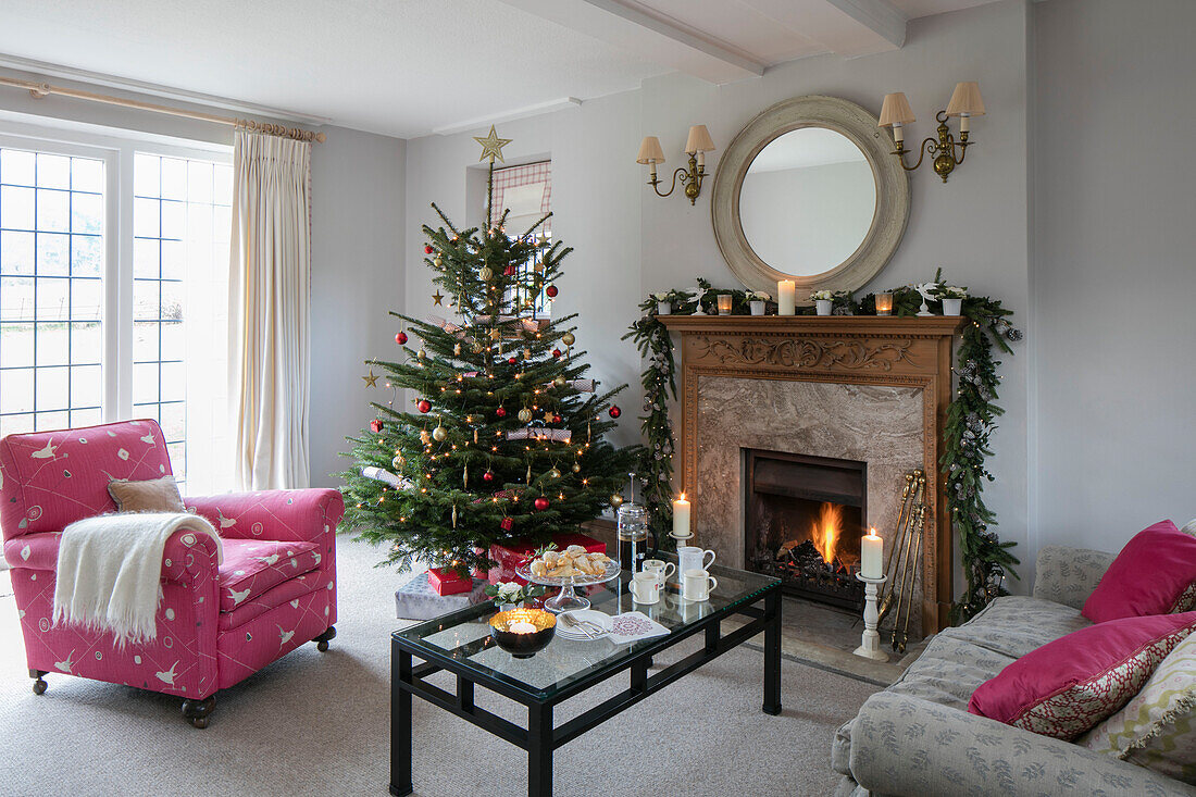 Rosafarbener Sessel und beleuchtetes Feuer mit Weihnachtsbaum am Fenster eines Hauses im Arts-and-Crafts-Stil in West Sussex, Großbritannien