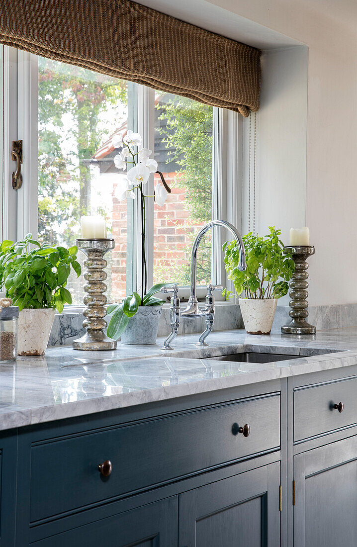 Large silver candlesticks and herbs at marble topped sink in window of Oxfordshire kitchen UK