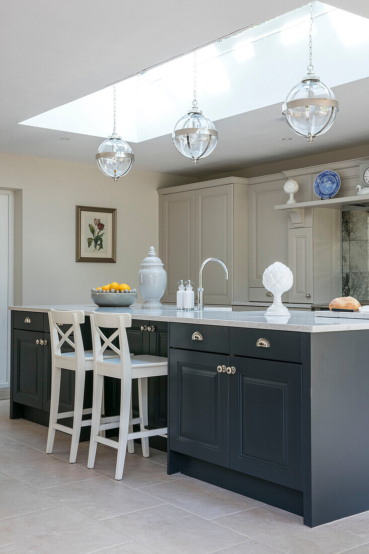 White bar stools at grey kitchen island under skylight with glass pendants in Sussex home UK
