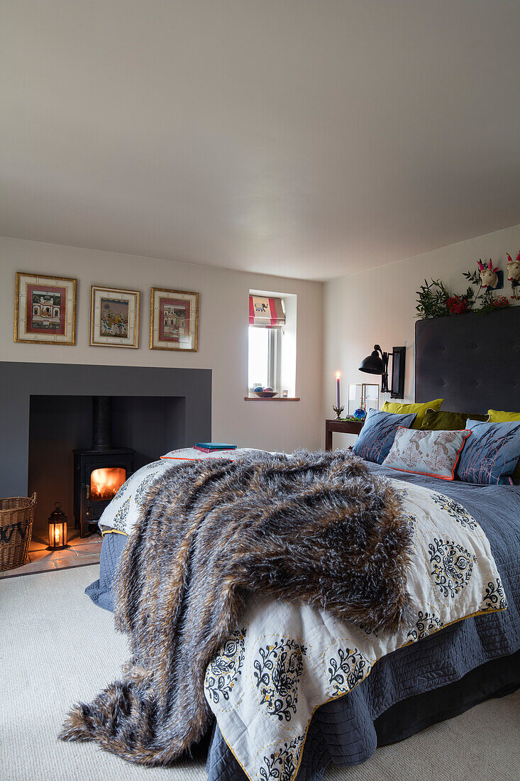 Faux fur and linen cushions on double bed with lit fire in Norfolk cottage England UK