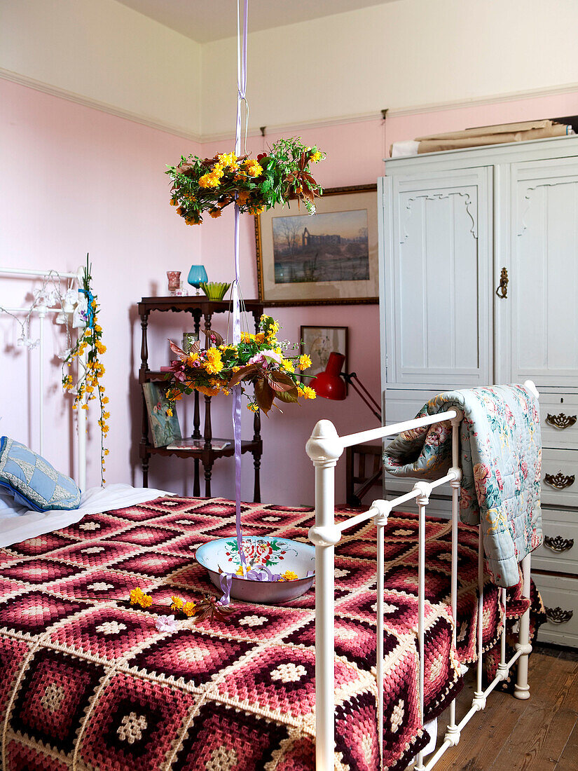Floral baskets hang on ribbon above metal bed with crochet blanket in Isle of Wight home UK