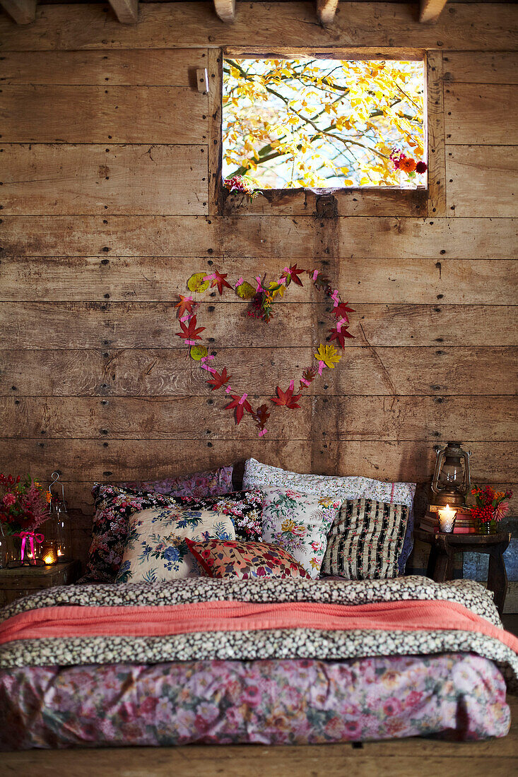 Verschiedene Blumenstoffe auf Matratze unter Blätterherz und Fenster in rustikaler Holzhütte Herbst UK