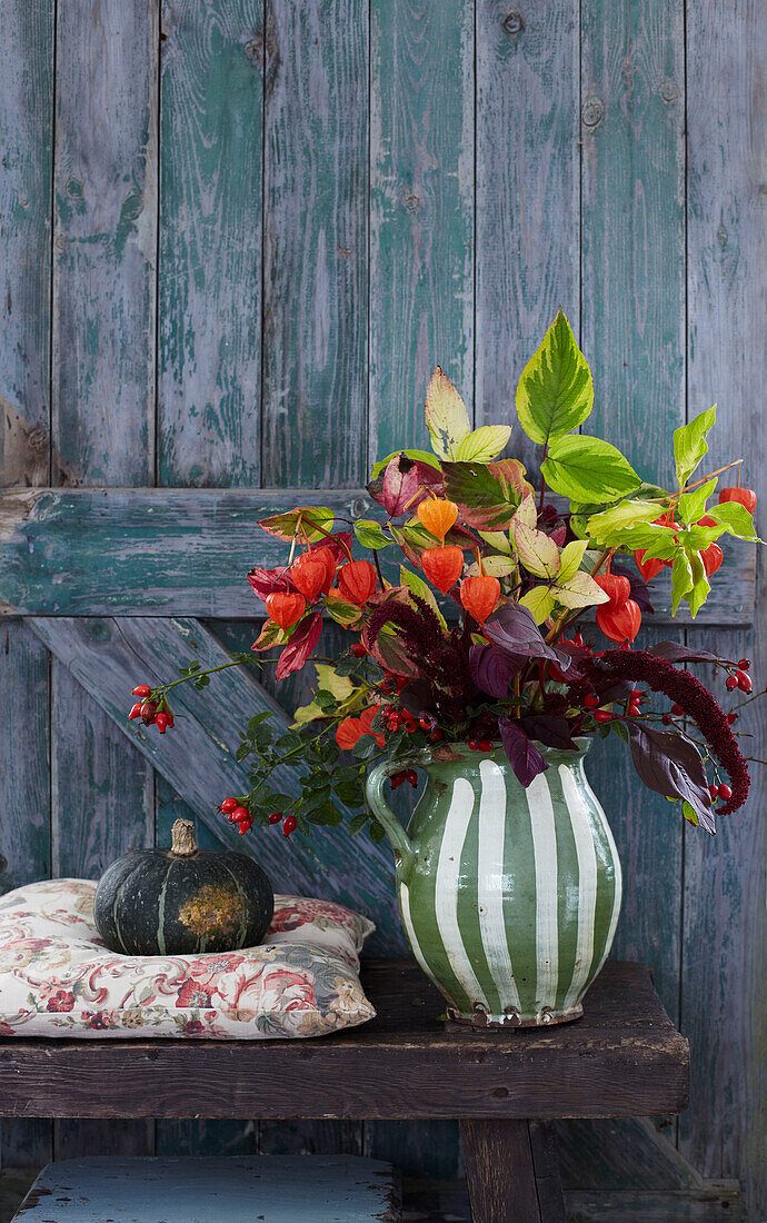 Rustic green door with wooden bench displaying an autumnal bouquet in a striped green and white jug with pumpkin on a cushion