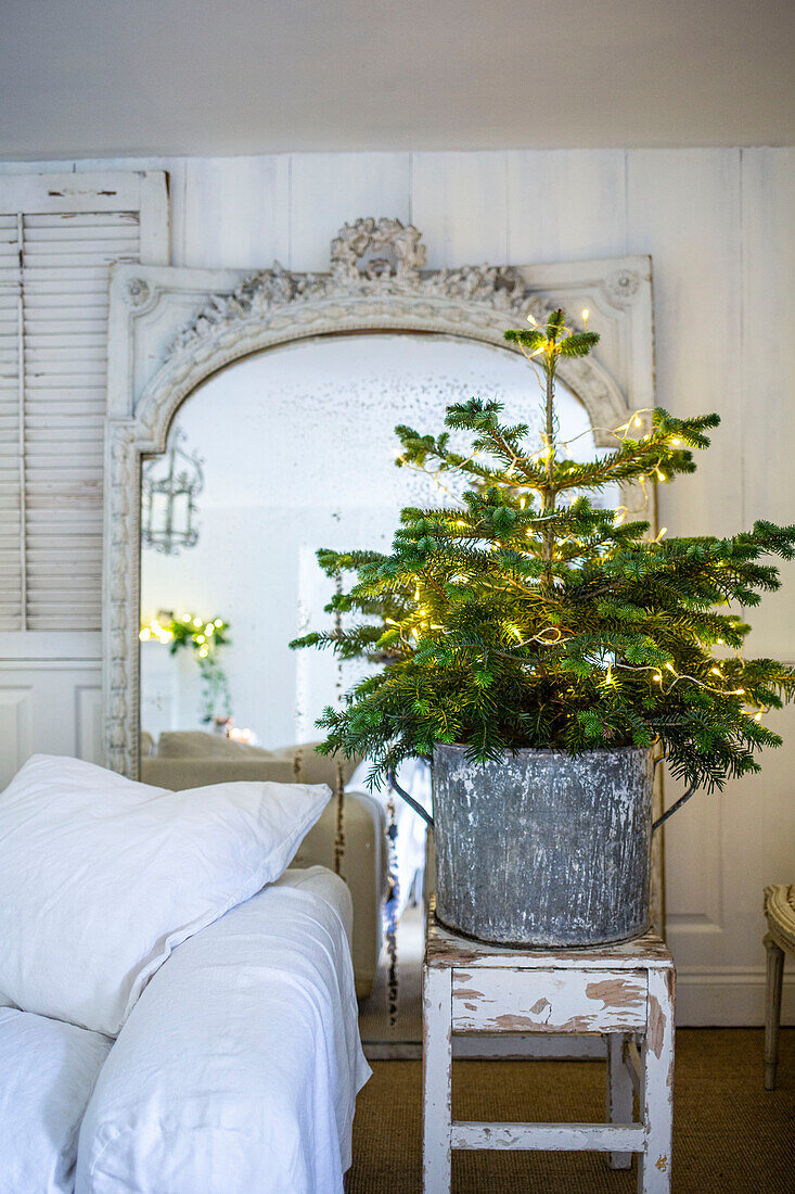 Scandi style living room with white loose covered sofa and small Christmas tree in galvanised planter and louvered shutters on the wall beside a large foxed mirror