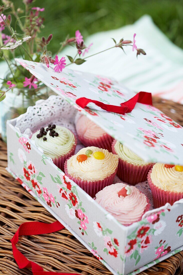 Box of pretty iced cupcakes in a floral covered cake box on a wicker hamper with a jar of wild flowers