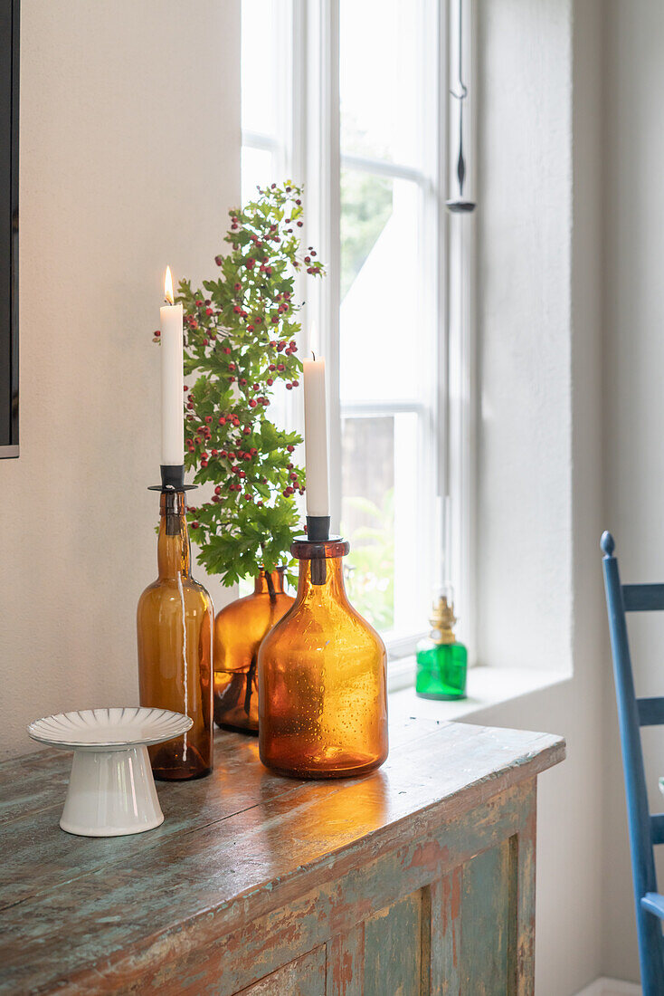 Candles in coloured glass bottles on wooden dresser next to window