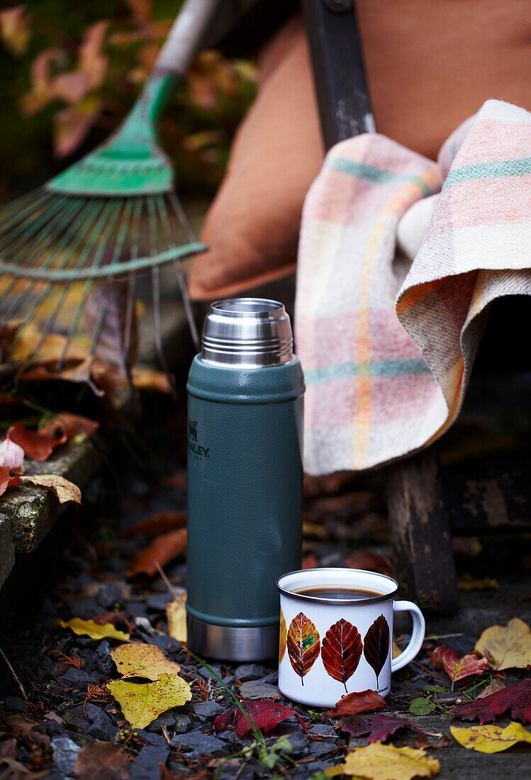 Thermos flask and cup next to leaf rake in autumn garden