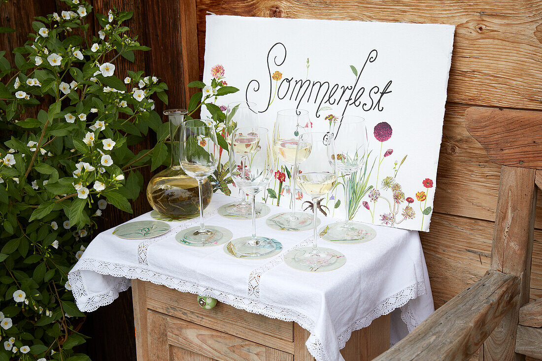 Small dresser with white tablecloth, wine glasses on coasters and sign Summer party