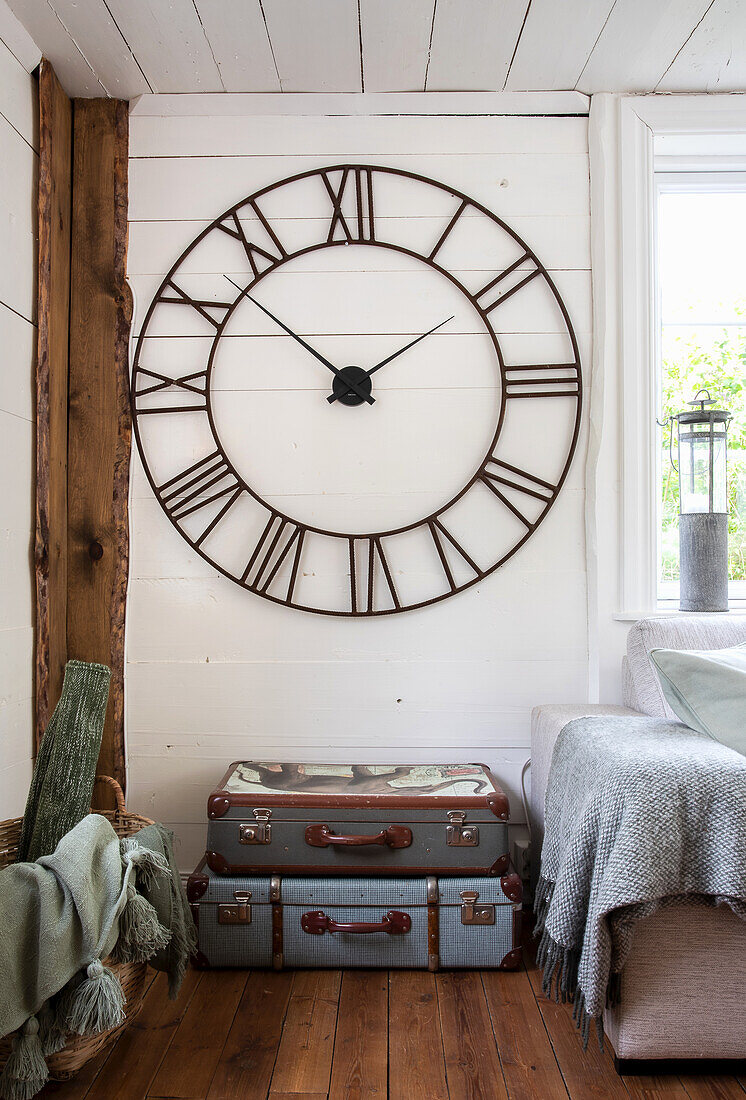 Large vintage-style wall clock above antique suitcases, corner sofa with blanket