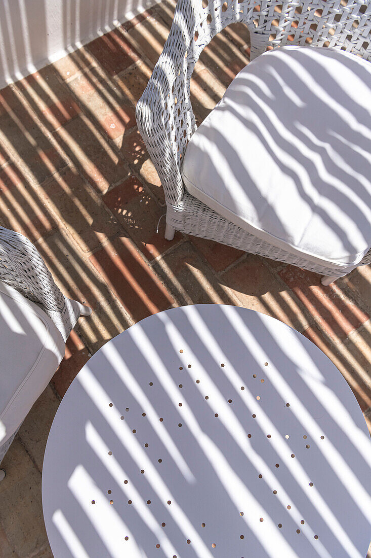 Weißer Gartentisch und Rattanstühle mit Polster auf Terrasse mit Schattenmuster