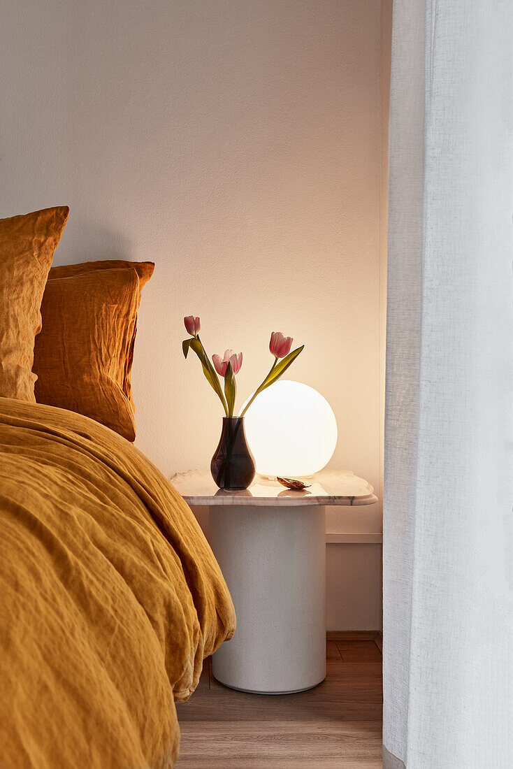 Apartment-Schlafzimmer mit Kurkumafarbener Bettwäsche, einem Beistelltisch aus Marmor, runder Lampe und Vase mit rosa Tulpen