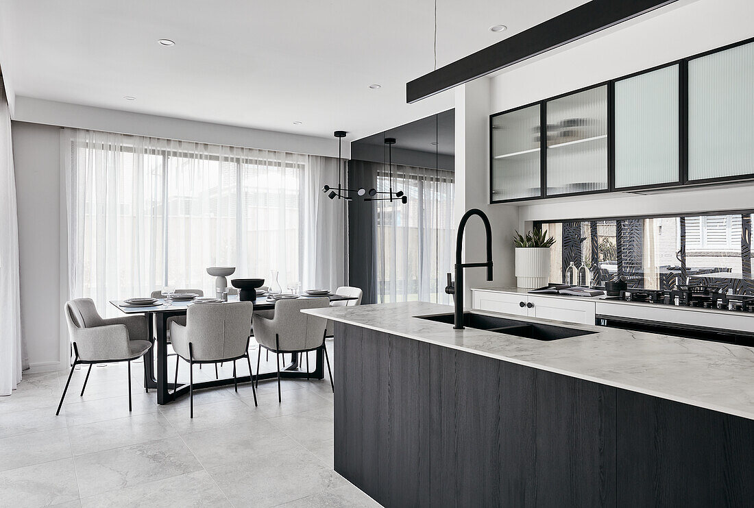 A modern monochromatic kitchen with a thin marble benchtop, black fittings and glass fronted cabinets with an open-plan dining room in the background