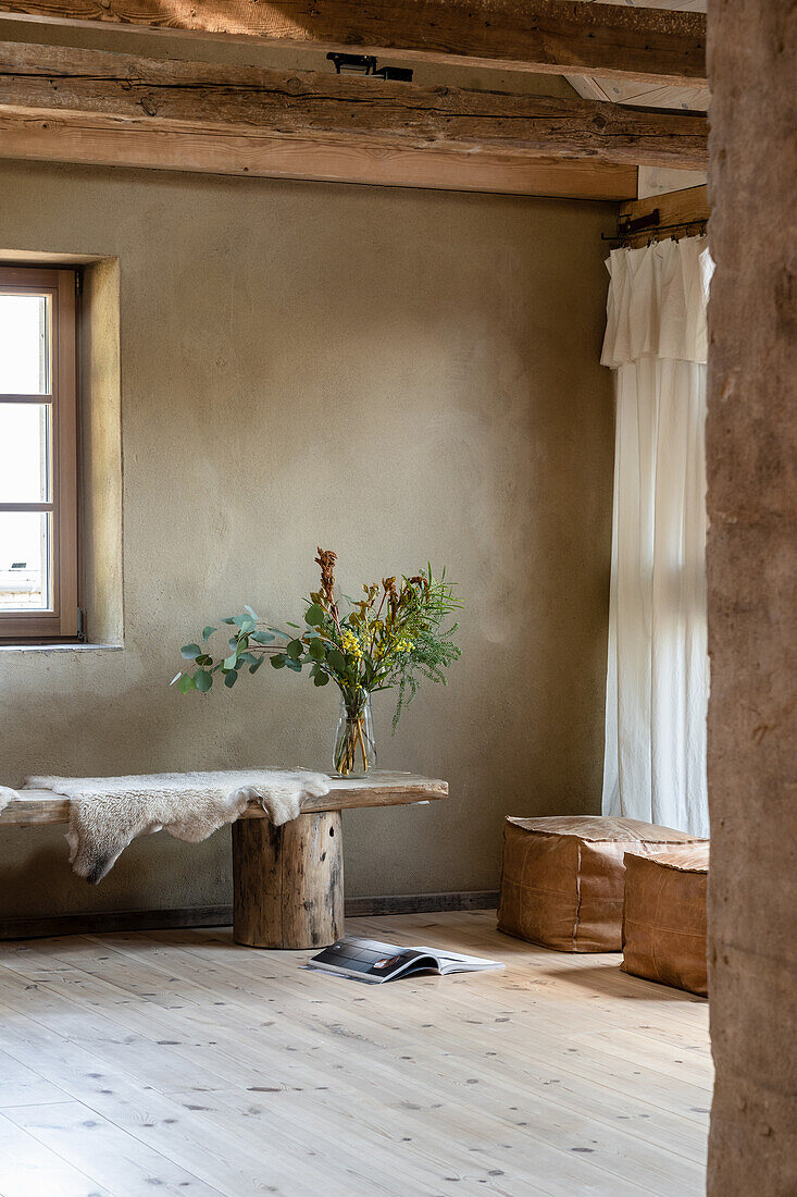 Wooden bench with sheepskin as table and leather square pouf in a room with clay plaster wall and wooden beams