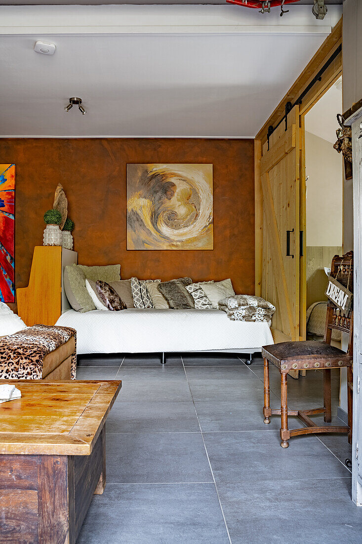 Tagesbett mit Kissensammlung vor rostbrauner Wand, im Vordergrund Vintage Stuhl und Tisch aus Holz
