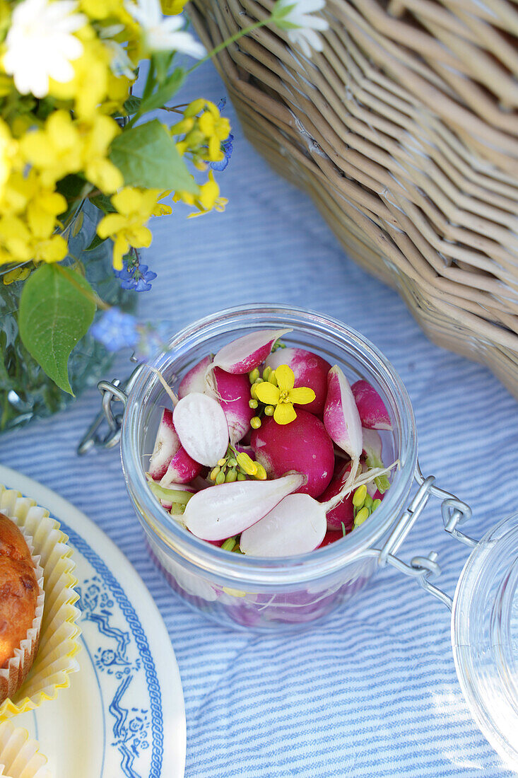 Glass jar with radishes next to picnic basket and flowers