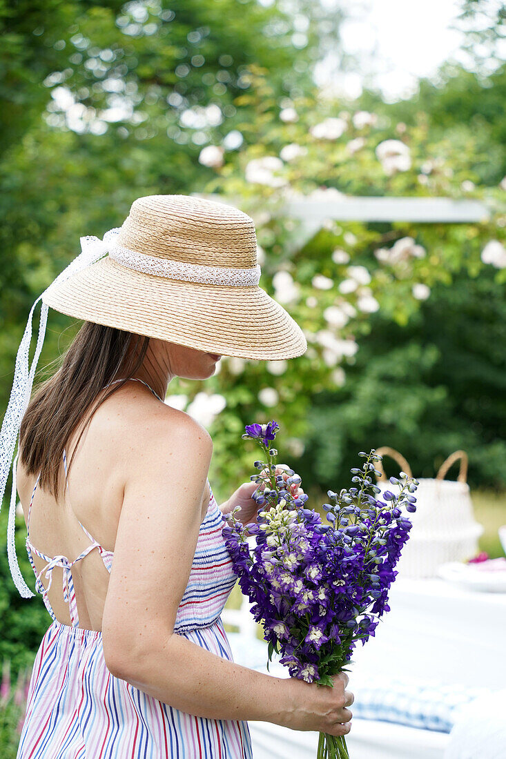 Frau im Sommerkleid mit Strohhut hält Rittersporn (Delphinium) im Garten