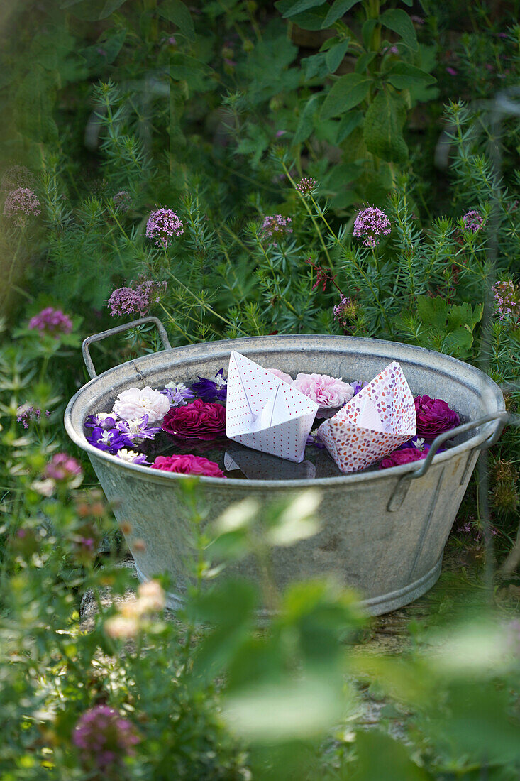 Blüten und Papierboote schwimmen in einer Zinkwanne im Garten