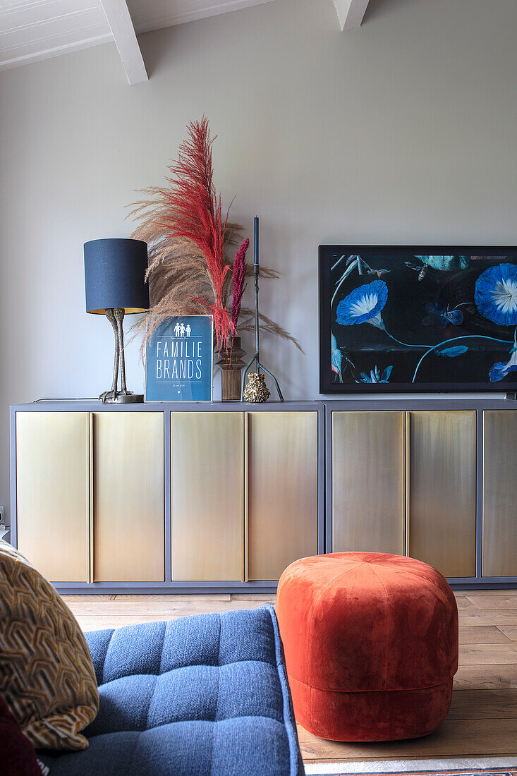 Wohnzimmer mit goldenem Sideboard, blauer Couch und rotem Hocker