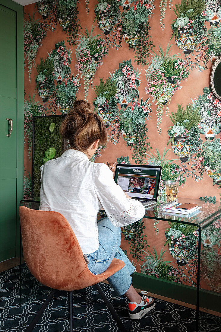 Frau sitzt im Home Office-Arbeitsplatz mit Glas-Schreibtisch und floraler Tapete