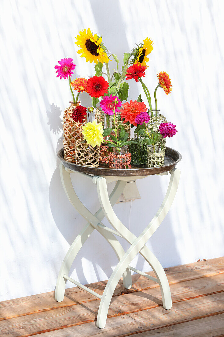 Glasvasen mit Sonnenblumen (Helianthus) und Gerbera auf kleinem Gartentisch