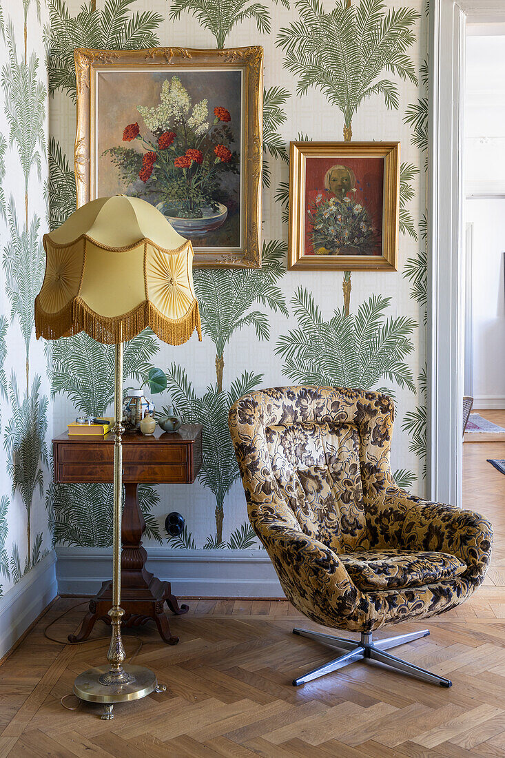 Reading armchair with baroque fabric upholstery, floor lamp and side table in front of palm pattern wallpaper