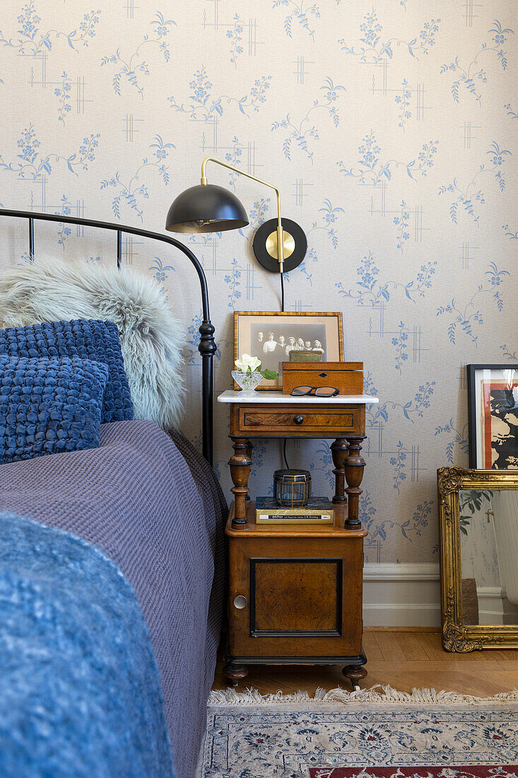 Antique wooden bedside table next to iron bed in front of blue, patterned wallpaper