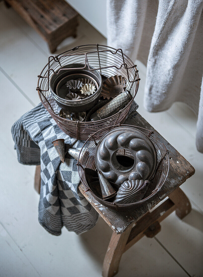 Wire basket with old baking tins on a rustic stool and patterned cloth