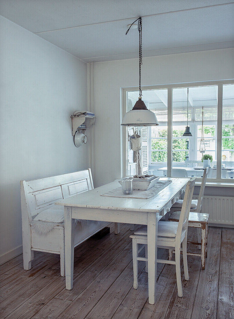 Dining area with white, rustic wooden table and bench in front of large windows