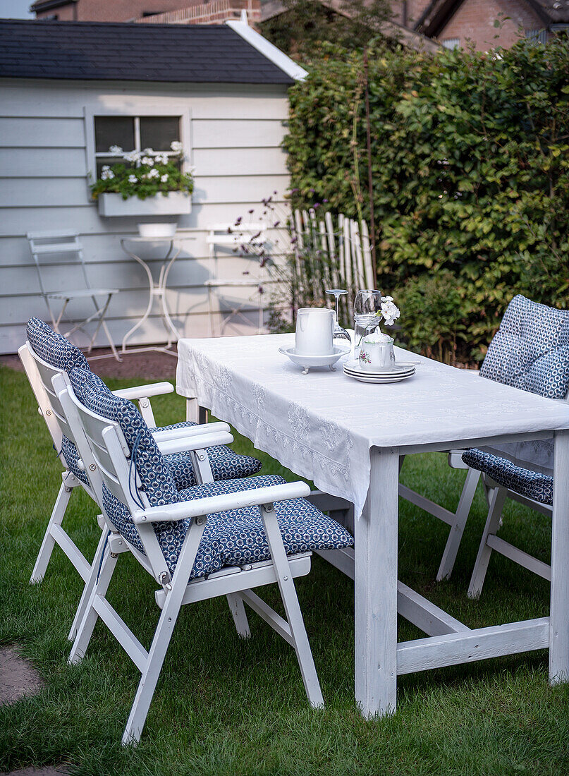 White garden table with blue cushioned chairs on lawn in front of garden shed