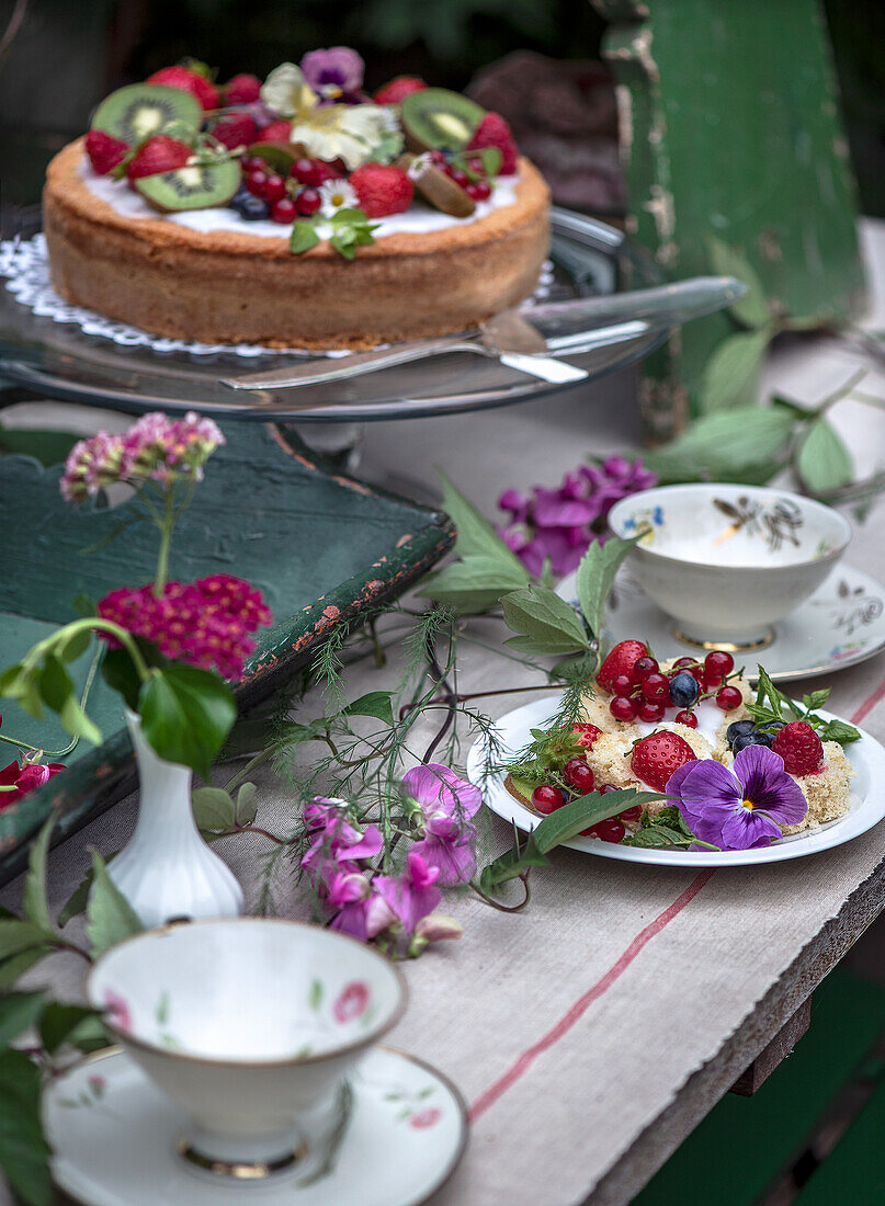 Sommerkuchen mit Beeren und Kiwi auf rustikal gedecktem Tisch mit Blumendekoration