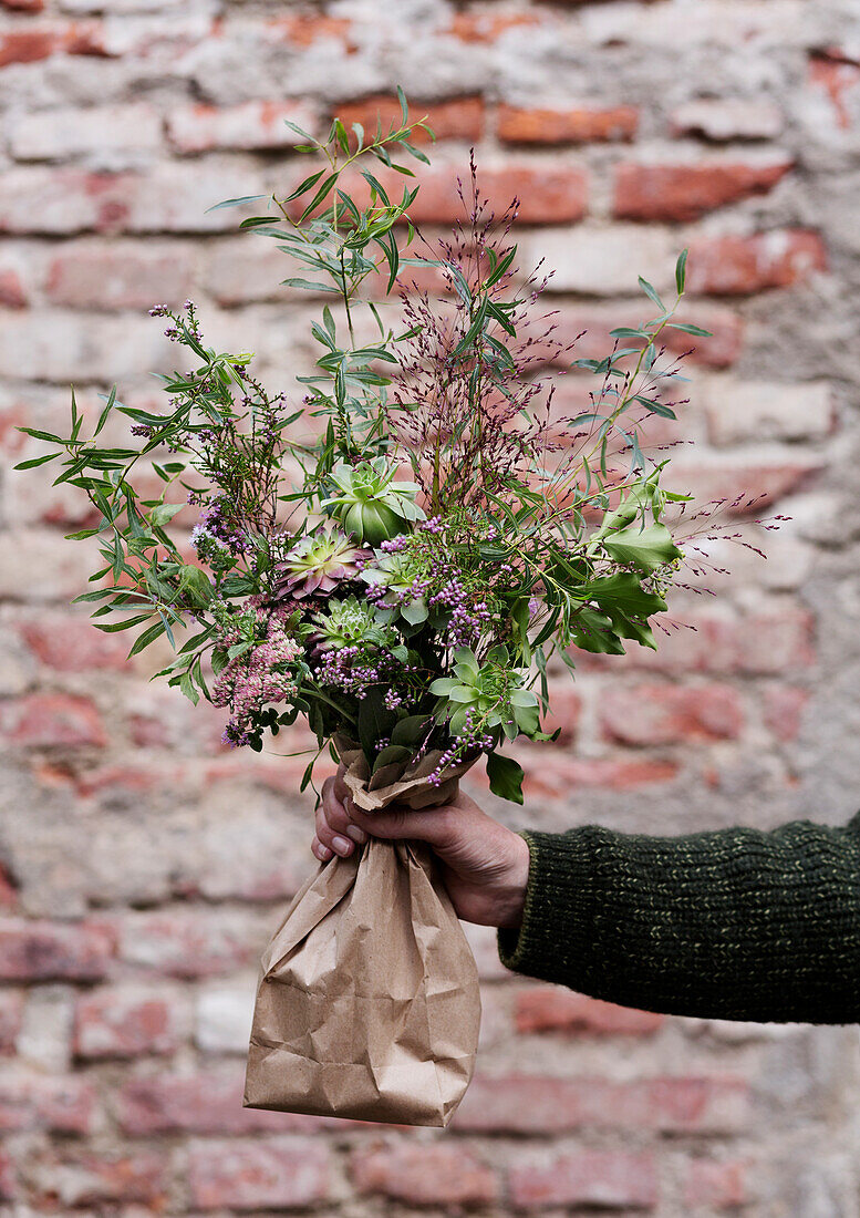 Hand holding bunch of wild flowers in brown paper in front of brick wall