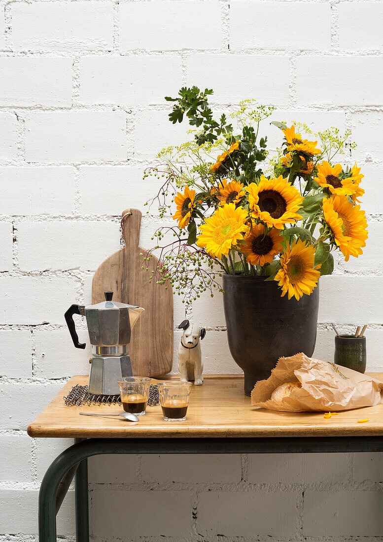 Holztisch mit Sonnenblumenstrauß in schwarzer Vase, Espressokocher und Tassen