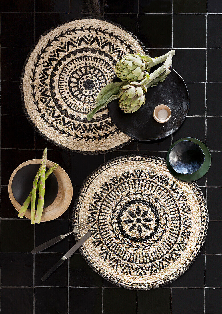 Black, tiled table surface with woven placemats, artichokes and asparagus