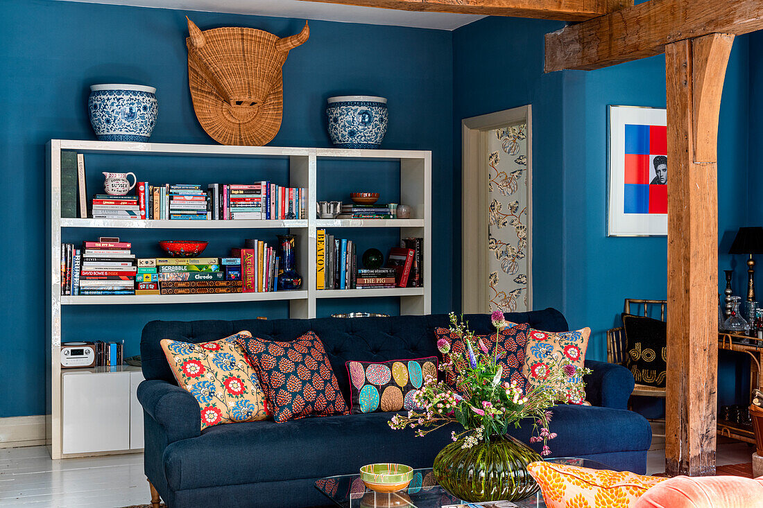 Living room with blue wall paint, bookshelf, dark blue sofa and decorative pillows in a colorful mix of patterns