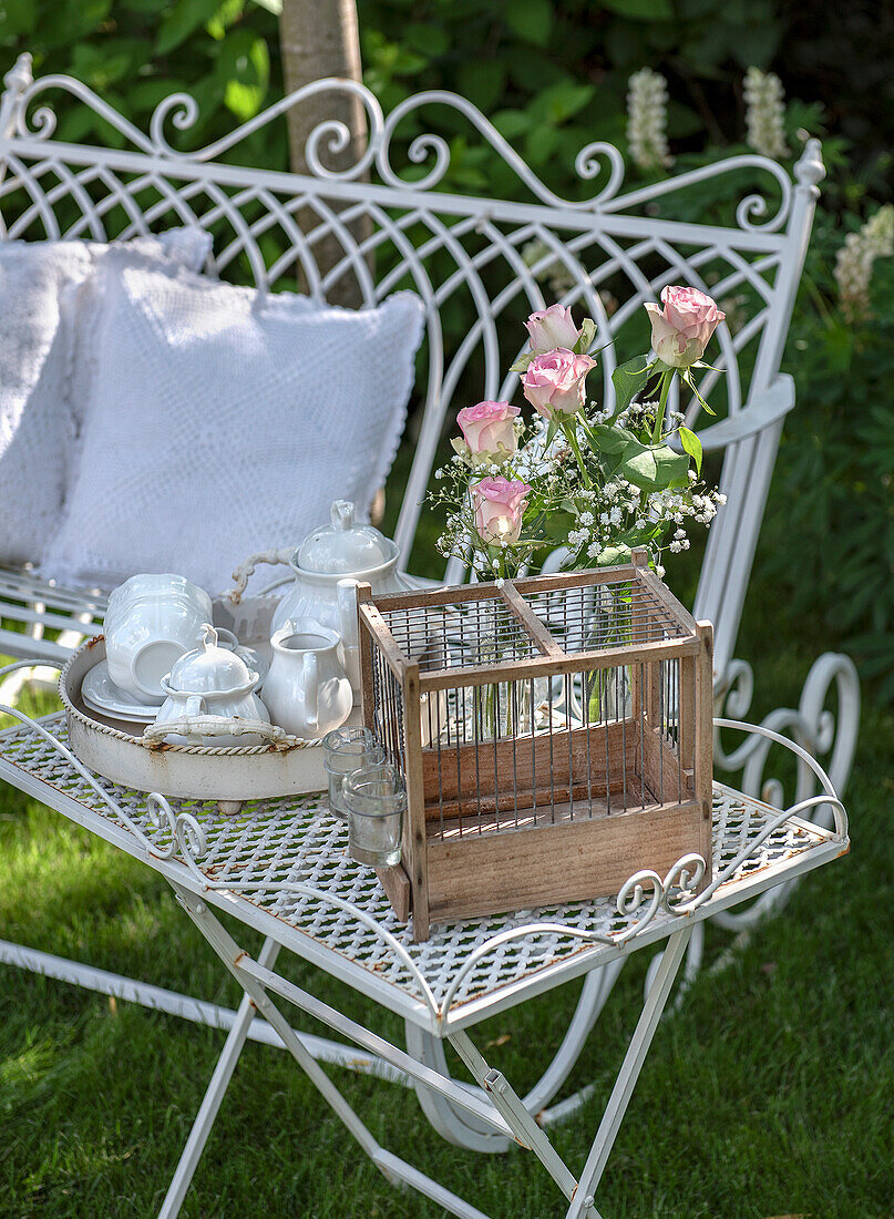 Romantic garden bench with tea service, bouquet of roses and birdcage on metal table