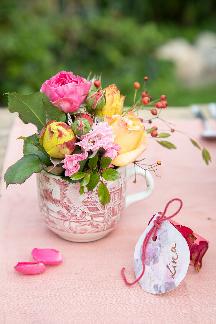 Flower arrangement with roses and rose hips in teacup on pink tablecloth